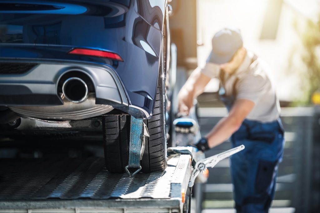 A delivery professional securing a blue car onto a flatbed transport truck using heavy-duty straps.