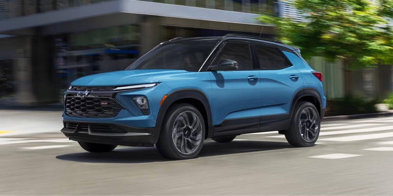 A bright blue Chevrolet Trailblazer RS with a black roof driving through a sunlit urban area with motion blur.