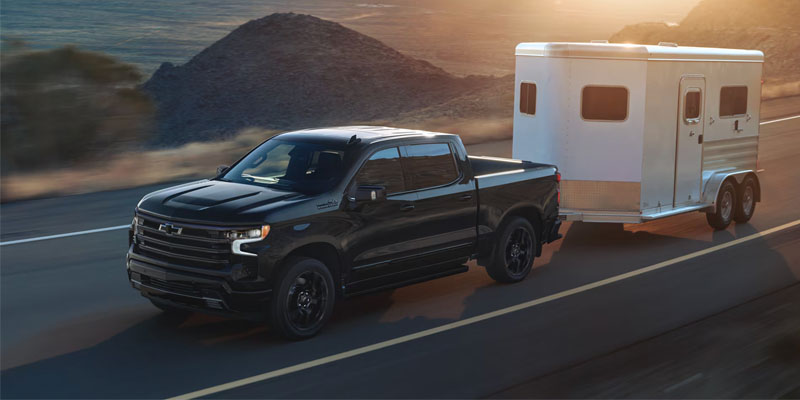 High-angle view of a black Chevrolet Silverado truck towing a white horse trailer down a desert highway at sunset.