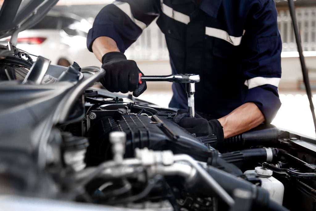 Close-up of a mechanic in black gloves using a socket wrench to work on a car engine under an open hood.
