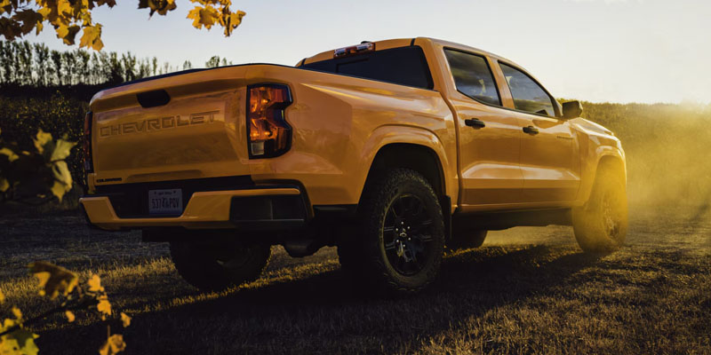 Rear view of a yellow Chevrolet Colorado pickup truck driving through a field during golden hour.