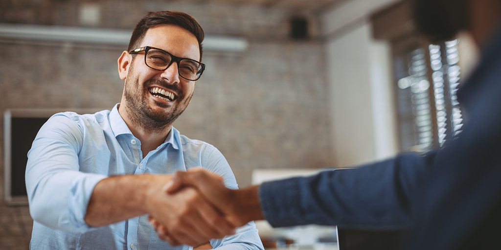 A smiling man with glasses and a light blue shirt shaking hands with someone across a table in an office setting.