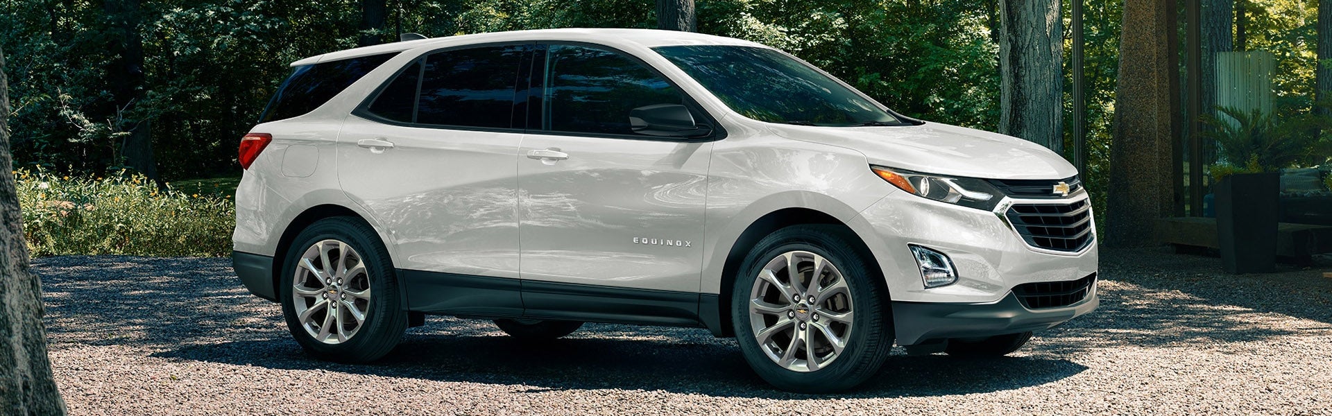A white pre-owned Chevrolet Equinox SUV parked on a gravel driveway surrounded by trees.