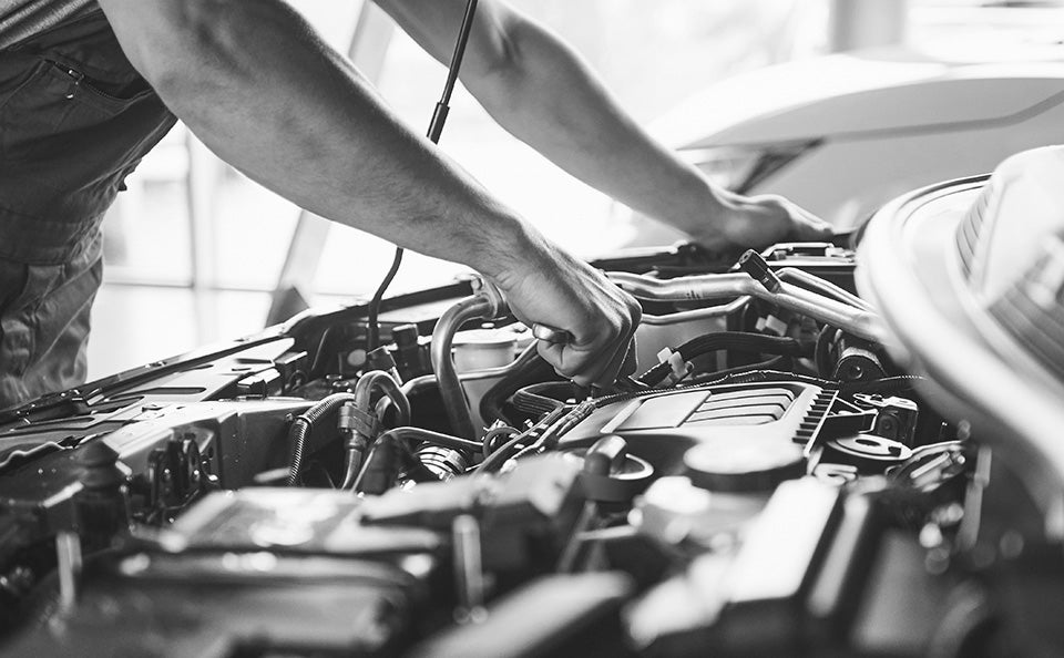 A black and white close-up of a mechanic working on a car engine under an open hood.